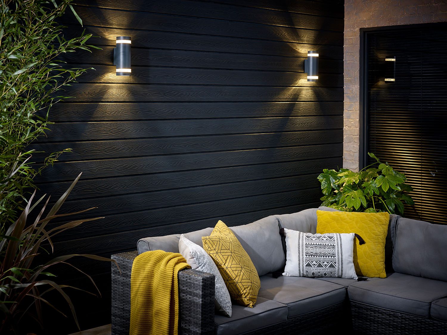 Black clad wall with two exterior lights behind a wicker sofa with yellow and grey cushions at night