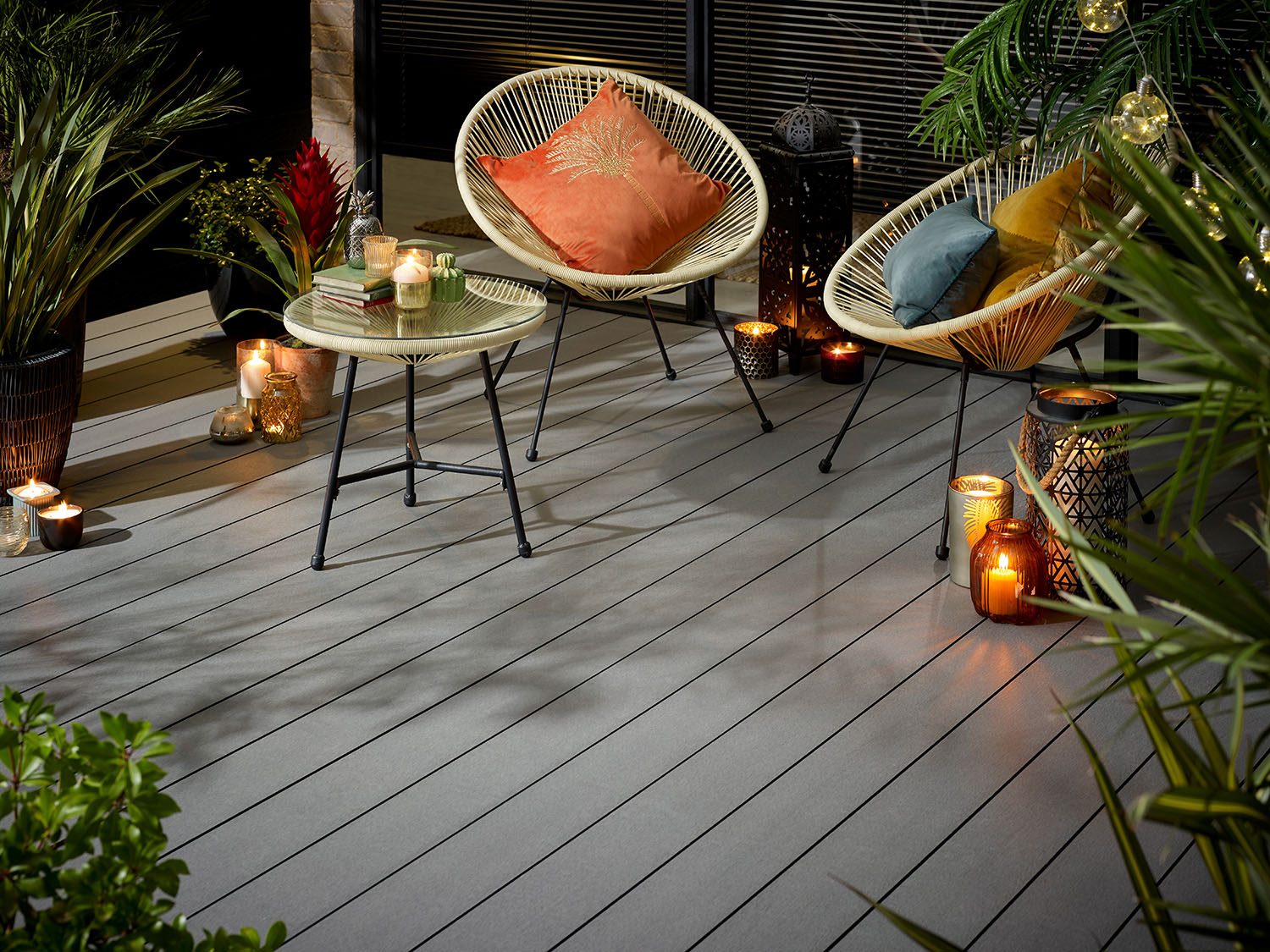 Grey wood grain decked courtyard with two cream chairs and table surrounded by plants at night with lit candles